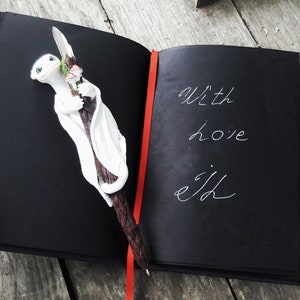 May include: A black journal with a red ribbon bookmark. The journal is open to a page with the words "With love, ejd" written in white ink. A white dragon figurine with pink flowers is on the left page.