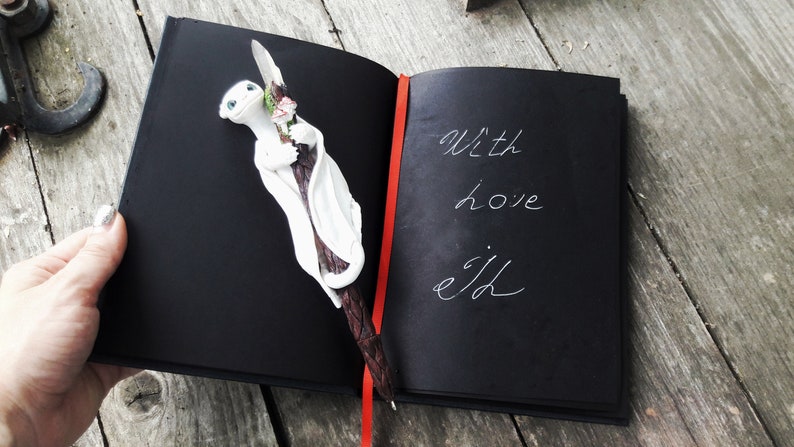 May include: A black journal with a red ribbon bookmark. The journal is open to a page with the words "With love, ejd" written in white ink. A white dragon figurine with pink flowers is on the left page.