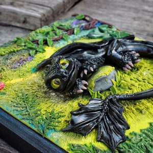 May include: A black dragon figurine with green eyes, lying on a green and yellow textured surface with a gray rock. The dragon has a black wing and a curled tail.