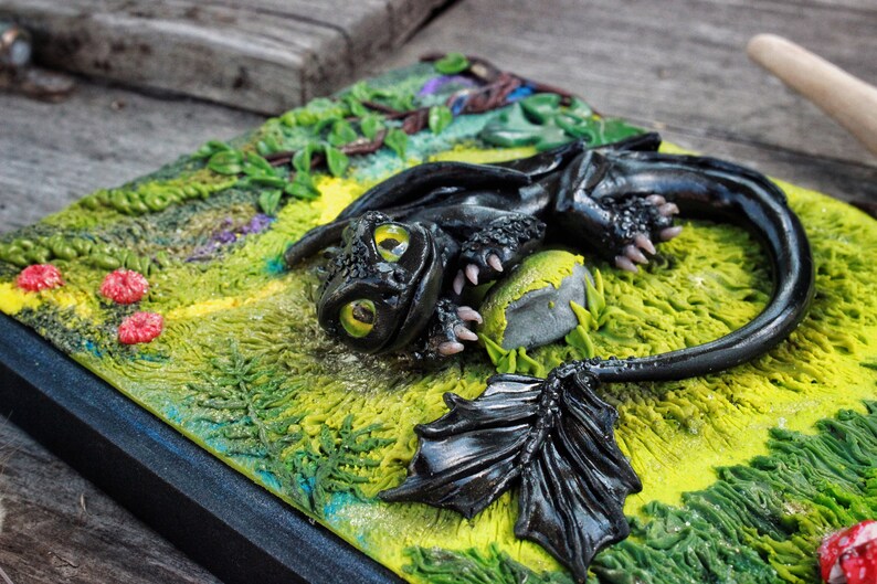 May include: A black dragon figurine with green eyes, lying on a green and yellow textured surface with a gray rock. The dragon has a black wing and a curled tail.