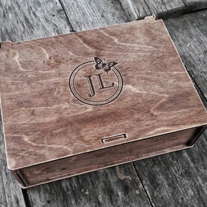 May include: A wooden box with a hinged lid. The lid is engraved with a circle containing the initials "JL" and a butterfly design.