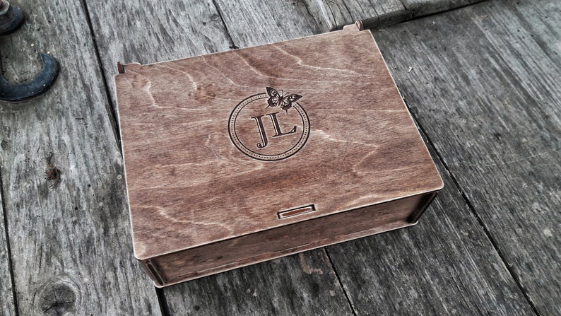 May include: A wooden box with a hinged lid. The lid is engraved with a circle containing the initials "JL" and a butterfly design.