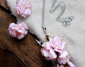 Puede incluir: Un conjunto de joyas con un collar y pendientes con colgantes de flores rosas y detalles dorados. El collar tiene una cadena plateada y los pendientes son tipo botón. El conjunto se exhibe sobre un paño de lino neutro sobre una superficie de madera.