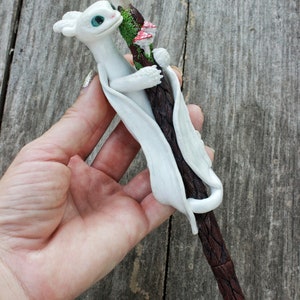 May include: White dragon figurine holding a brown and white pen with a clear crystal at the top. The pen has green moss and red mushrooms on it.