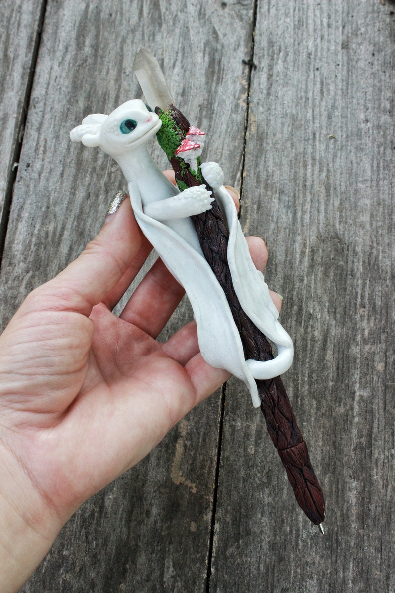 May include: White dragon figurine holding a brown and white pen with a clear crystal at the top. The pen has green moss and red mushrooms on it.