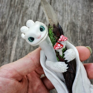 May include: White dragon figurine holding a crystal wand with red and white mushrooms. The dragon is curled around the wand and has a friendly expression.