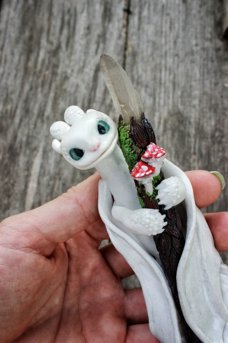 May include: White dragon figurine holding a crystal wand with red and white mushrooms. The dragon is curled around the wand and has a friendly expression.