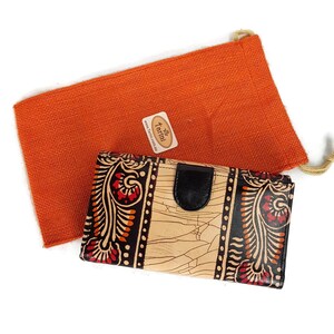 Könnte beinhalten: Eine beige Ledergeldbörse mit einem schwarzen und roten Blumenmuster. Die Geldbörse hat eine schwarze Lederklappe mit einem Druckknopfverschluss. Die Geldbörse ist in einer kleinen orangefarbenen Jutesack mit einem Kordelzugverschluss verpackt. Der Sack hat ein braunes Etikett mit dem Text "Tarini" und einer Website-Adresse.