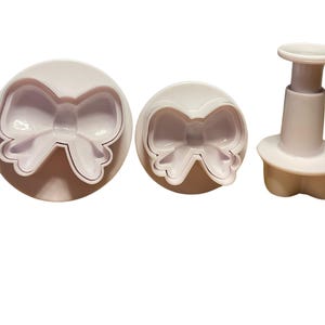 May include: Three plastic cookie cutters in the shape of a bow. The cutters are white and have a smooth finish. The largest cutter is a single piece, while the other two are two-piece cutters with a plunger.