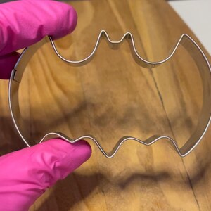 May include: A silver metal cookie cutter shaped like a bat. The cutter is held in a pink gloved hand.