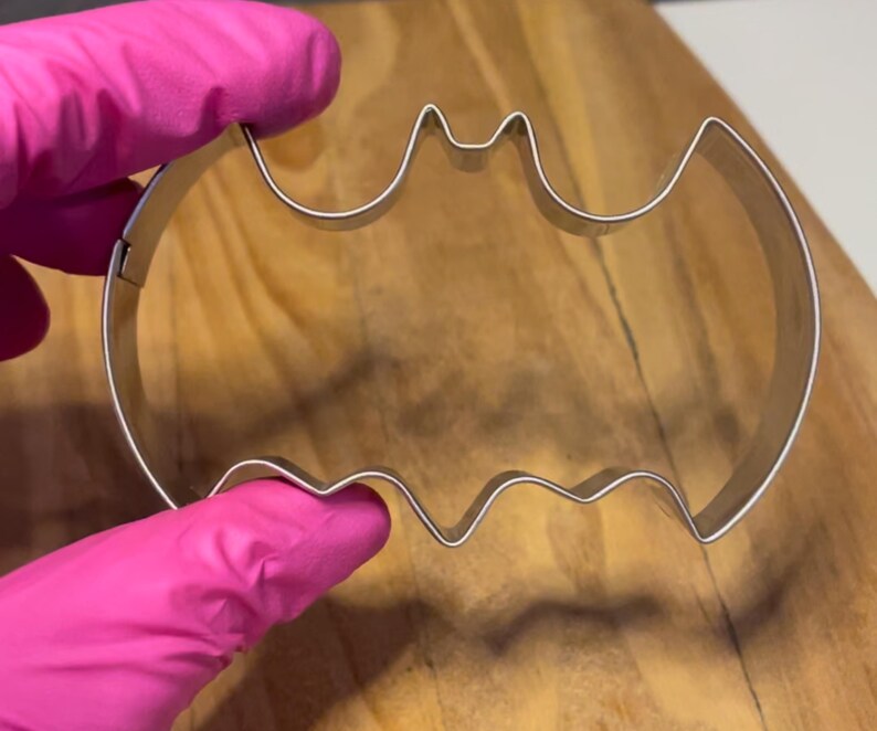 May include: A silver metal cookie cutter shaped like a bat. The cutter is held in a pink gloved hand.