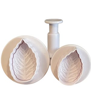 May include: Two white plastic leaf-shaped cookie cutters with a plunger. The larger cutter has a detailed leaf design.