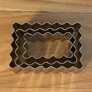 May include: Three silver metal cookie cutters in the shape of a rectangle with scalloped edges. The cutters are nested inside each other, with the largest cutter on the bottom and the smallest cutter on top.