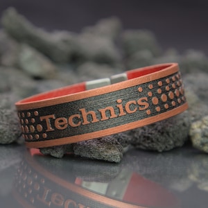 Puede incluir: Una pulsera negra y cobre con la palabra "Technics" grabada en ella. La pulsera tiene un forro interior rojo y un patrón de pequeños círculos.
