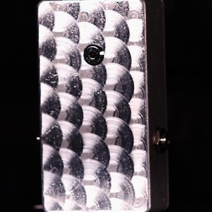 May include: A silver guitar effects pedal with a textured, scale-like pattern on the front. The pedal has a black button and a silver casing. The design is a close-up shot against a dark background.