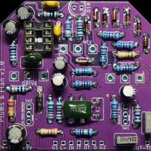 May include: A purple printed circuit board labelled Plums & Flan v1.0 featuring various electronic components including resistors with colour-coded bands, electrolytic capacitors, a black integrated circuit socket, and diodes soldered onto the surface.