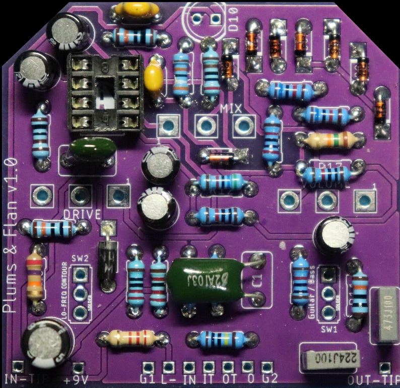 May include: A purple printed circuit board labelled Plums & Flan v1.0 featuring various electronic components including resistors with colour-coded bands, electrolytic capacitors, a black integrated circuit socket, and diodes soldered onto the surface.