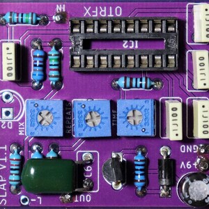 May include: A purple printed circuit board labeled SLAP v1.1 and OTRFX featuring a black integrated circuit socket, three blue potentiometers labeled MIX, REPEATS, and TIME, several blue resistors, white capacitors, a green capacitor, and a black diode.