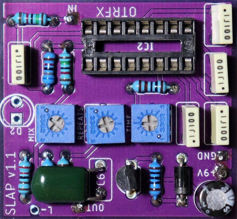 May include: A purple printed circuit board labeled SLAP v1.1 and OTRFX featuring a black integrated circuit socket, three blue potentiometers labeled MIX, REPEATS, and TIME, several blue resistors, white capacitors, a green capacitor, and a black diode.