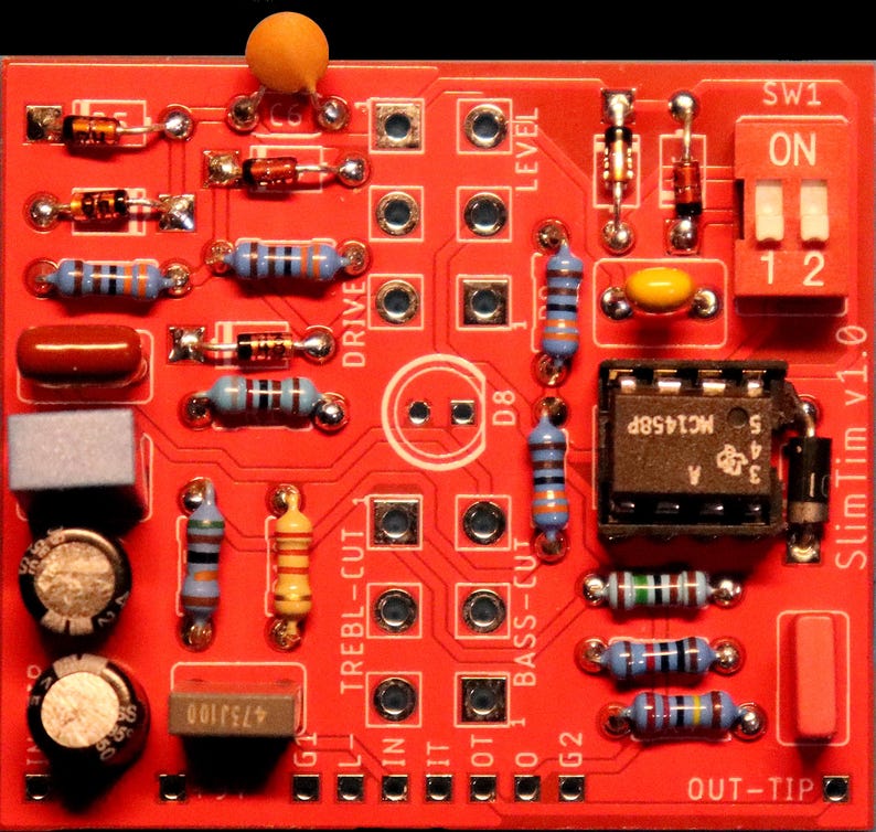 Peut inclure: Une carte de circuit imprim&eacute; rouge marqu&eacute;e SlimTim v1.0 comprenant divers composants &eacute;lectroniques, notamment des r&eacute;sistances bleues, jaunes et brunes, une puce de circuit int&eacute;gr&eacute; noire marqu&eacute;e MC1458P, un interrupteur blanc et des condensateurs.