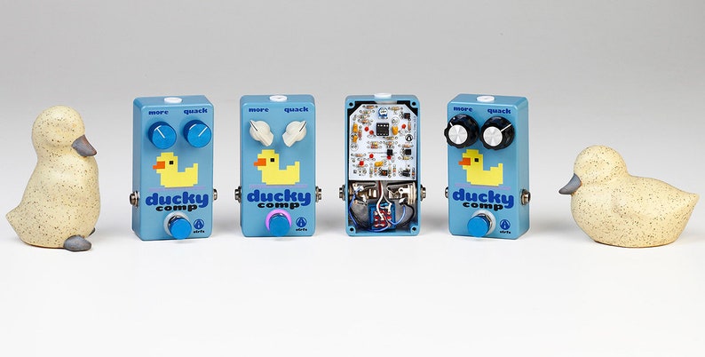 May include: Three light blue rectangular guitar effect pedals labelled Ducky Comp with pixelated yellow duck graphics, blue, white, and black knobs, and one pedal with an exposed circuit board, flanked by two speckled beige duck figurines.