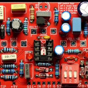May include: A red printed circuit board labelled OmniRat v1.0 featuring various electronic components including resistors, capacitors, a black integrated circuit socket, two red LEDs, and a three-position DIP switch labelled with a key for settings.
