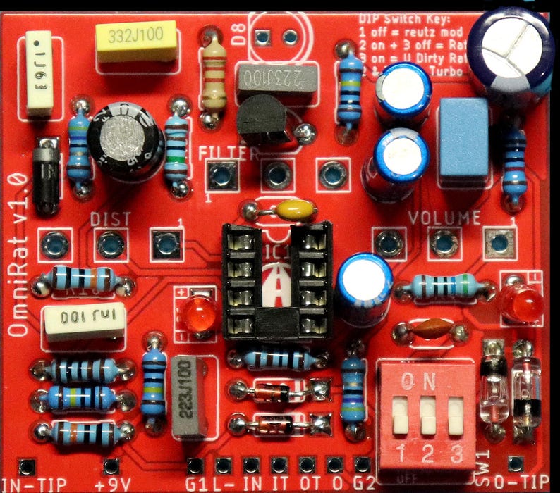 May include: A red printed circuit board labelled OmniRat v1.0 featuring various electronic components including resistors, capacitors, a black integrated circuit socket, two red LEDs, and a three-position DIP switch labelled with a key for settings.