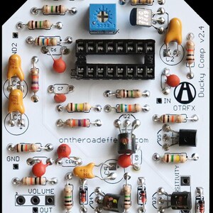 May include: A white printed circuit board labelled Ducky Comp v2.4 and ontheroadeffects.com. The board features a black integrated circuit socket, a blue potentiometer, various tan resistors with coloured bands, orange capacitors, and black transistors.