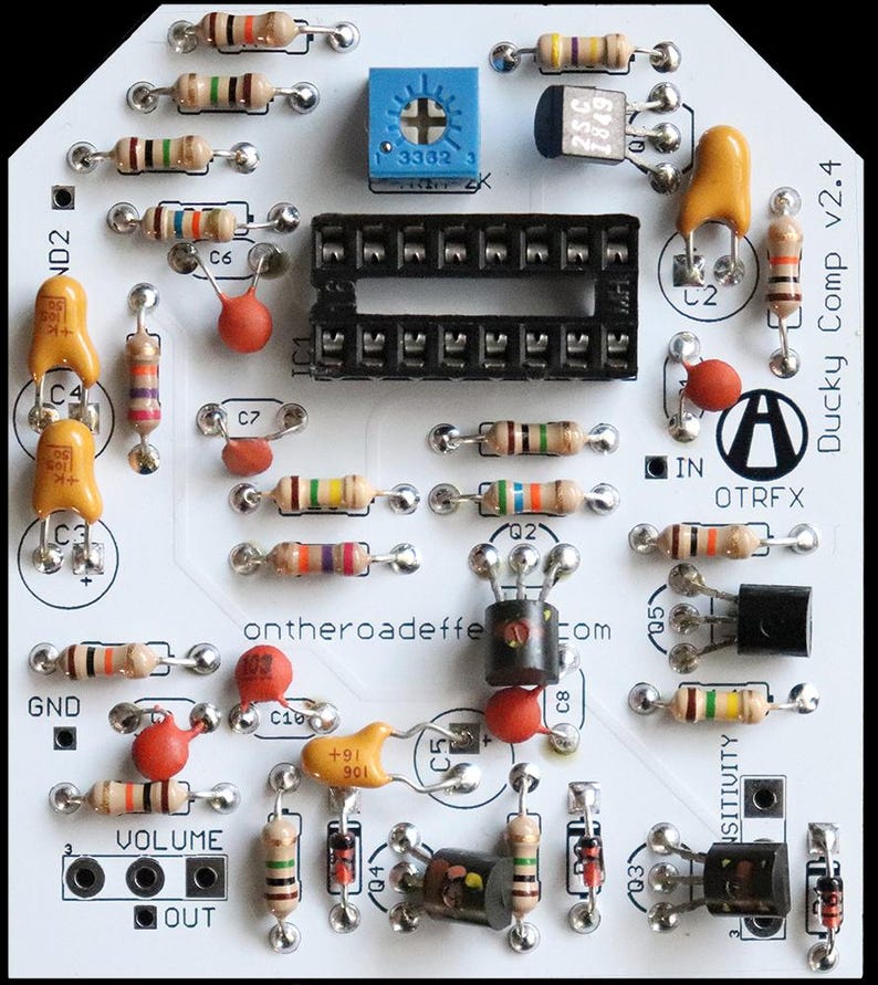 May include: A white printed circuit board labelled Ducky Comp v2.4 and ontheroadeffects.com. The board features a black integrated circuit socket, a blue potentiometer, various tan resistors with coloured bands, orange capacitors, and black transistors.
