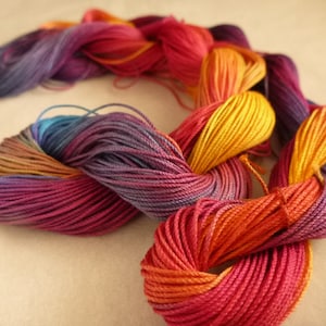 May include: A vibrant skein of yarn in a mix of colours including purple, orange, yellow, and red. The yarn is tightly wound and looks soft, suitable for knitting or crochet. The colours blend, creating a gradient effect.