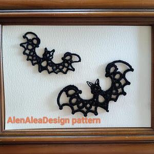 Op de afbeelding: Twee zwarte kanten vleermuisdecoraties in een houten lijst. De vleermuizen hebben gedetailleerde opengewerkte vleugels en lichamen. De tekst "AlenAleaDesign pattern" is zichtbaar. De lijst is warmbruin.