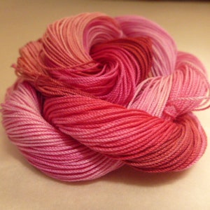 Puede incluir: Un ovillo de hilo en tonos rosas y rojos. El hilo está bien enrollado, con una textura suave y un degradado de colores que va del rosa claro al rojo intenso. El enrollado ordenado sugiere su uso en proyectos de manualidades.