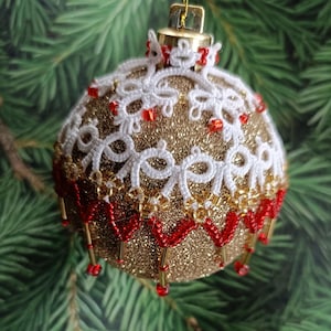 May include: A gold glitter Christmas bauble with white lace and red bead detailing. The bauble has a gold cap and is set against a green fir tree. The round bauble features a festive design.