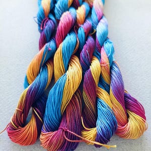 May include: A bundle of six colourful embroidery floss strands, each with a gradient of orange, yellow, blue, and purple.