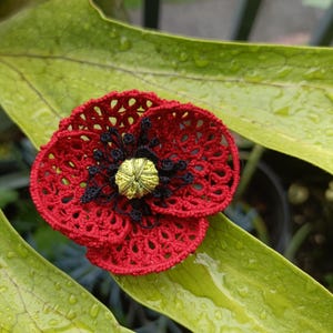 Könnte beinhalten: Eine handgefertigte Häkelblume in Rot, Schwarz und Gelb. Die Blume hat ein durchbrochenes Design mit einem strukturierten Zentrum. Sie liegt auf einem großen, grünen Blatt mit Wassertropfen. Dieser Dekorationsartikel ist wahrscheinlich eine Brosche oder ein Schmuckstück.