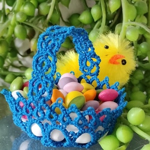 Può includere: Un cestino di crochet blu pieno di pezzi di cioccolato ricoperti di caramelle colorate. Un pulcino giallo con un becco rosso spunta da dietro il cestino.