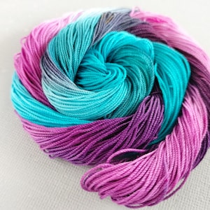 May include: A skein of colourful embroidery floss in shades of pink, purple, teal, and blue. The floss is wound into a tight coil.