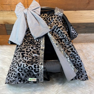 May include: A gray and black leopard print car seat cover with silver glitter accents. The cover has a matching gray bow on top.