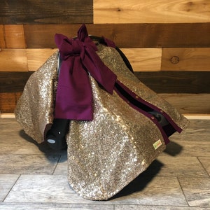 May include: Gold sequin car seat cover with a purple bow. The cover has a zipper on the side and a small tag that says "Little Lovely."