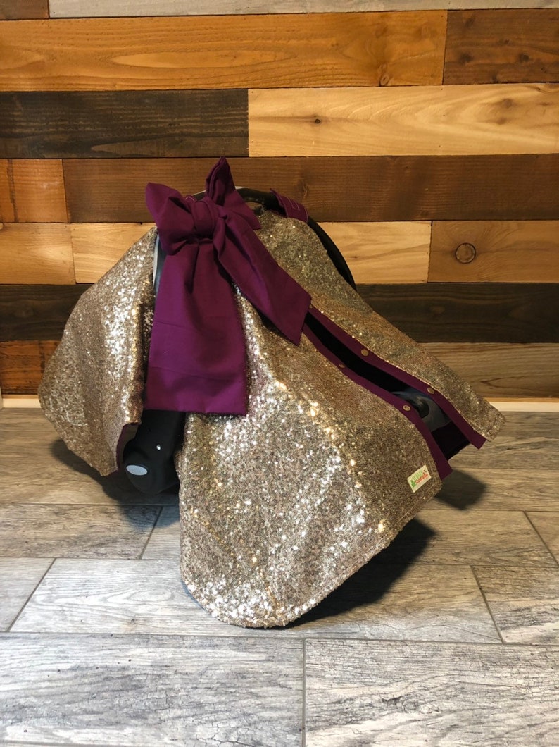 May include: Gold sequin car seat cover with a purple bow. The cover has a zipper on the side and a small tag that says "Little Lovely."