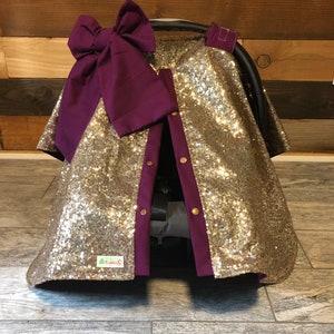 May include: Gold sequin car seat cover with a purple bow and purple trim. The cover has a zipper closure and a small green label that says "Cozy Covers".