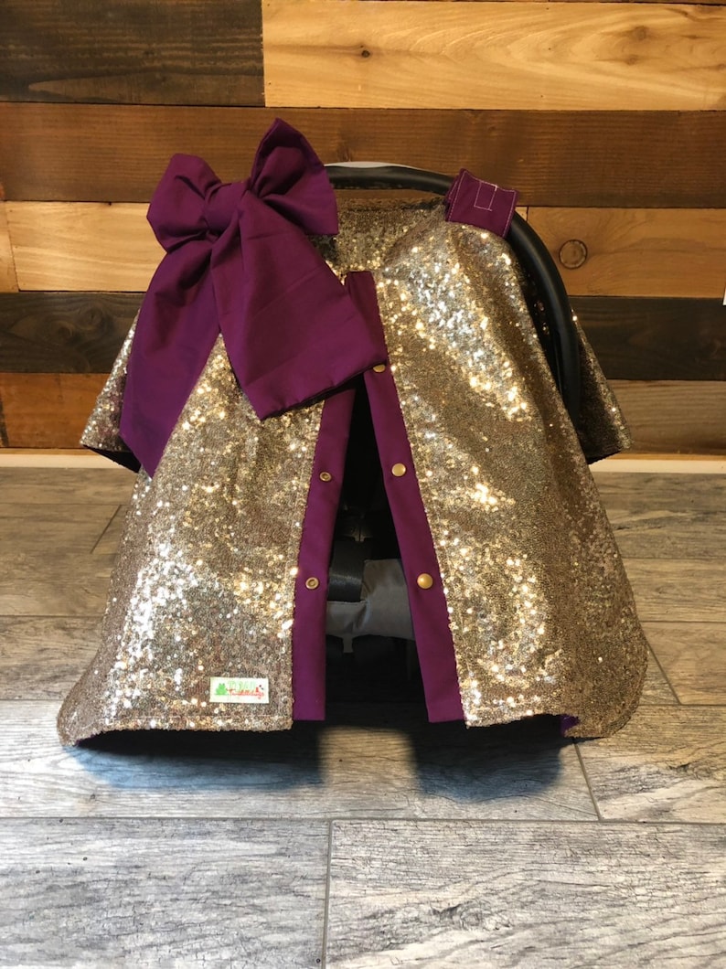 May include: Gold sequin car seat cover with a purple bow and purple trim. The cover has a zipper closure and a small green label that says "Cozy Covers".