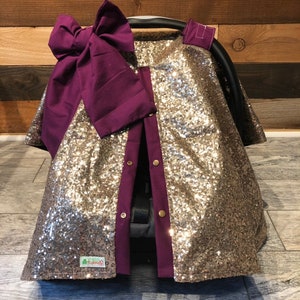 May include: Gold sequin car seat cover with a purple bow and purple trim. The cover has buttons down the front and a small green label with the word "Little" on it.