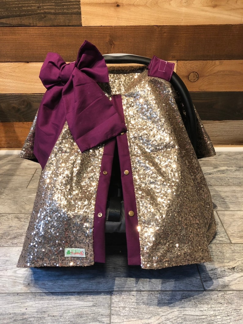 May include: Gold sequin car seat cover with a purple bow and purple trim. The cover has buttons down the front and a small green label with the word "Little" on it.
