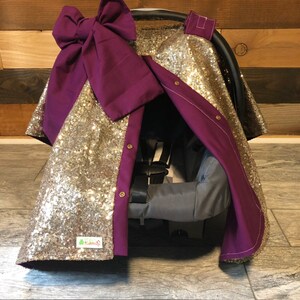 May include: Gold sequin car seat cover with a purple bow and a purple lining. The cover has buttons down the side and a small tag that says "Kisses & Cuddles".