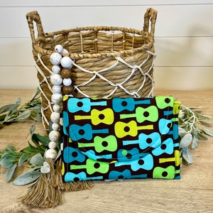 May include: A brown fabric pouch with a repeating pattern of blue and green guitars. The pouch is folded in half and sitting on a wooden surface. A woven basket with a white beaded garland is in the background.
