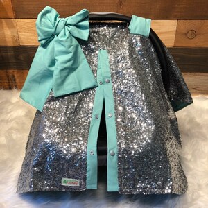 May include: Silver sequin car seat cover with a light blue bow and trim. The cover has snaps down the front and a small green label that says "Little Luxuries".