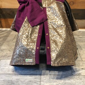 May include: Gold sequin car seat cover with a purple bow and a purple trim. The cover has a button closure and a small green label with the text "Little Twigs".