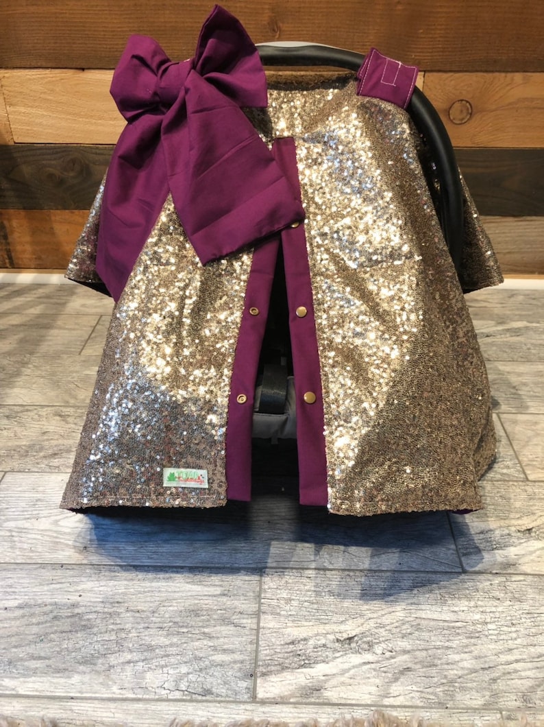 May include: Gold sequin car seat cover with a purple bow and a purple trim. The cover has a button closure and a small green label with the text "Little Twigs".