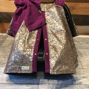 May include: Gold sequin car seat cover with a purple bow and buttons. The cover has a split down the back for easy access to the car seat straps.